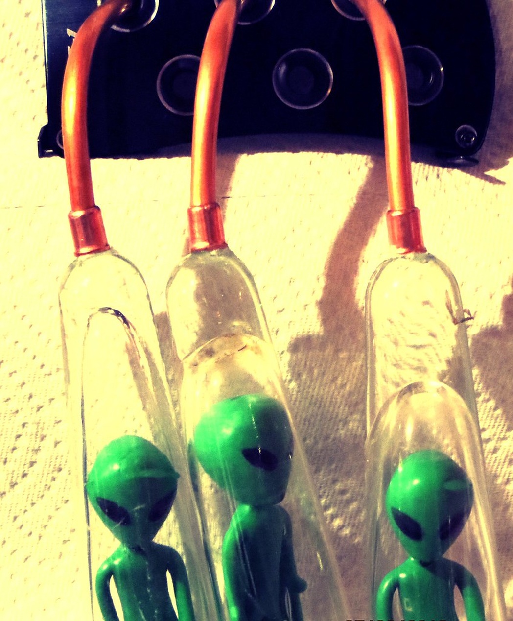 A close-up photograph of novelty items showing green alien figurines inside glass tubes with copper-colored tops. The alien figures have the stereotypical large heads, black almond-shaped eyes, and slender bodies associated with pop culture depictions of extraterrestrials. This appears to be a decorative or novelty item rather than scientific content about UFO radiation effects.
